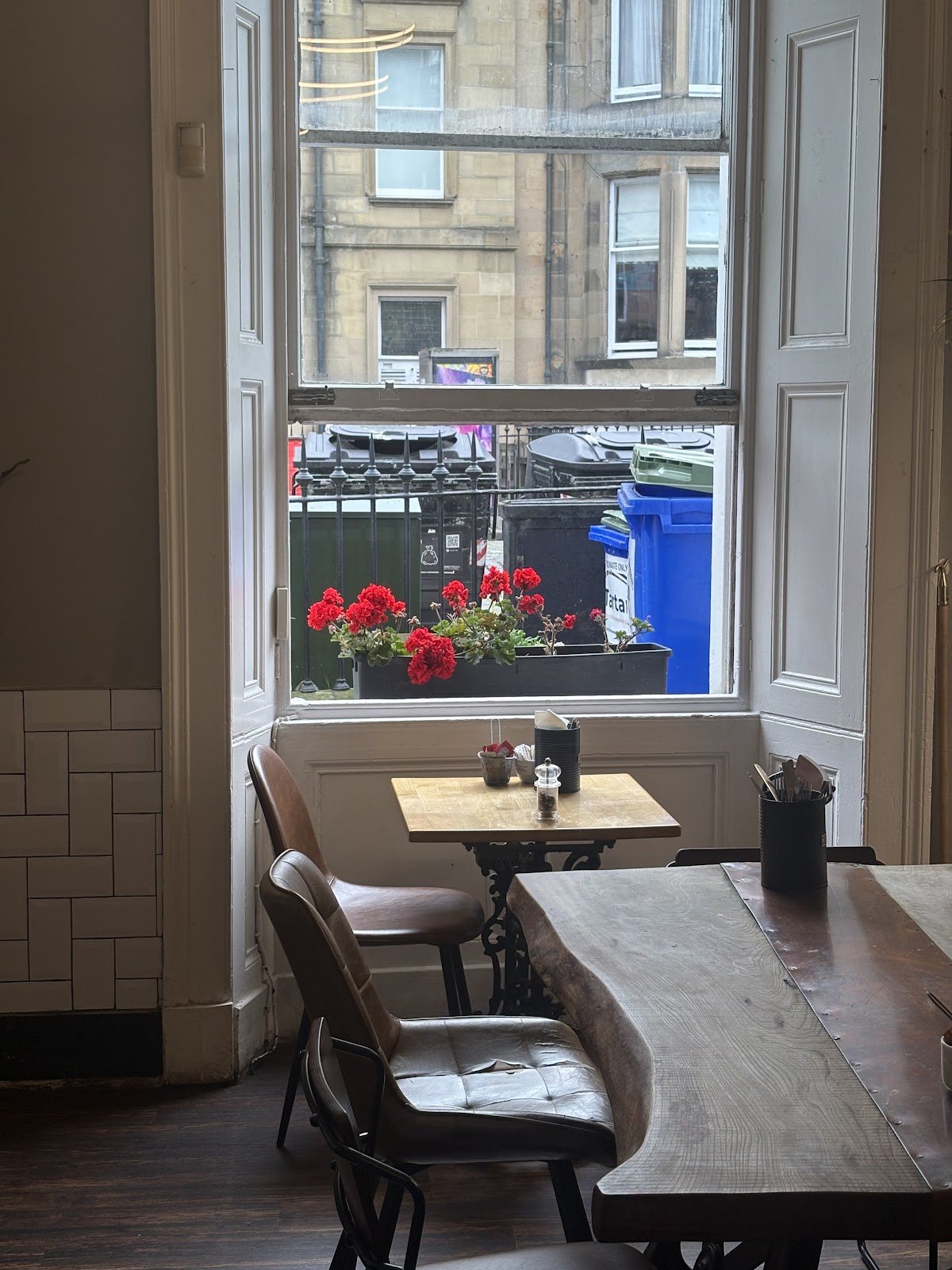 A table by the tall sash window, white shutters folded back and a window box of red geraniums outside, looking onto the sandstone tenements across Haymarket Terrace.