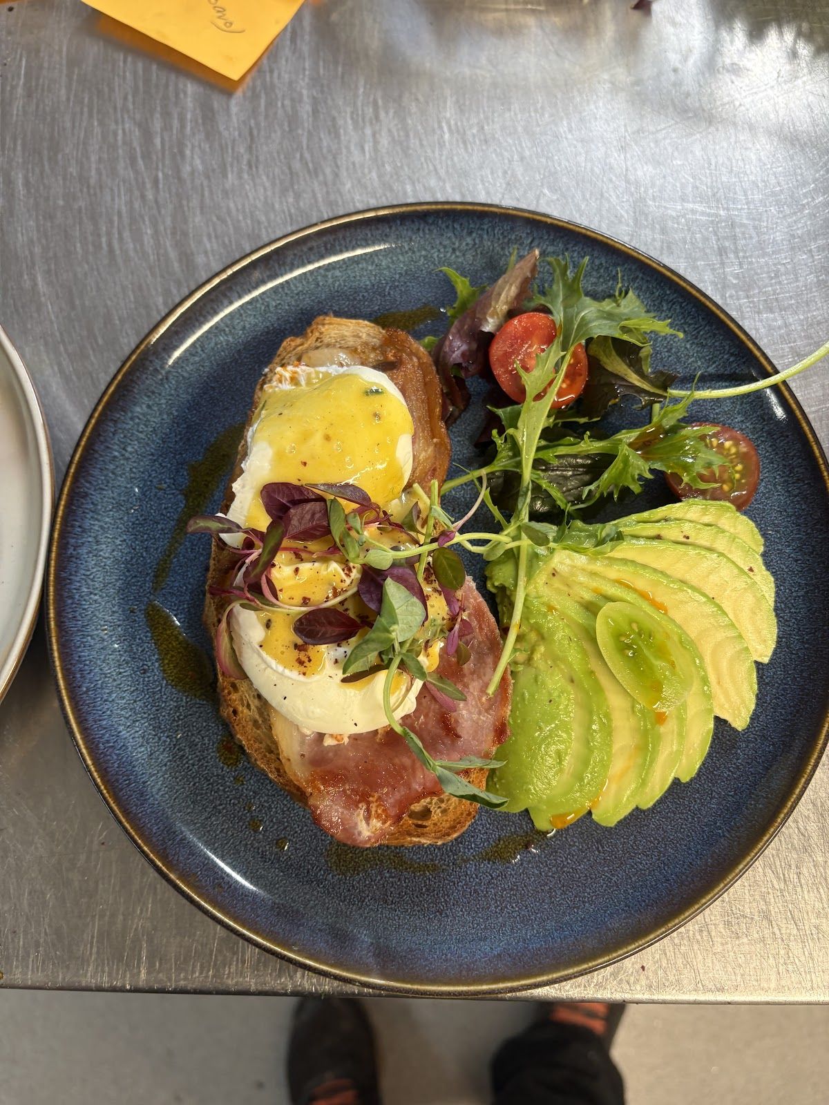 Poached eggs with hollandaise and bacon on toasted sourdough, fanned avocado, micro herbs and cherry tomatoes on a navy stoneware plate.