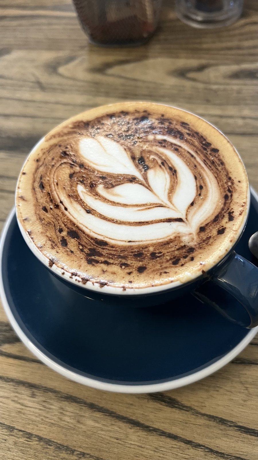 A cappuccino with a rosetta in a navy cup on a wooden table.