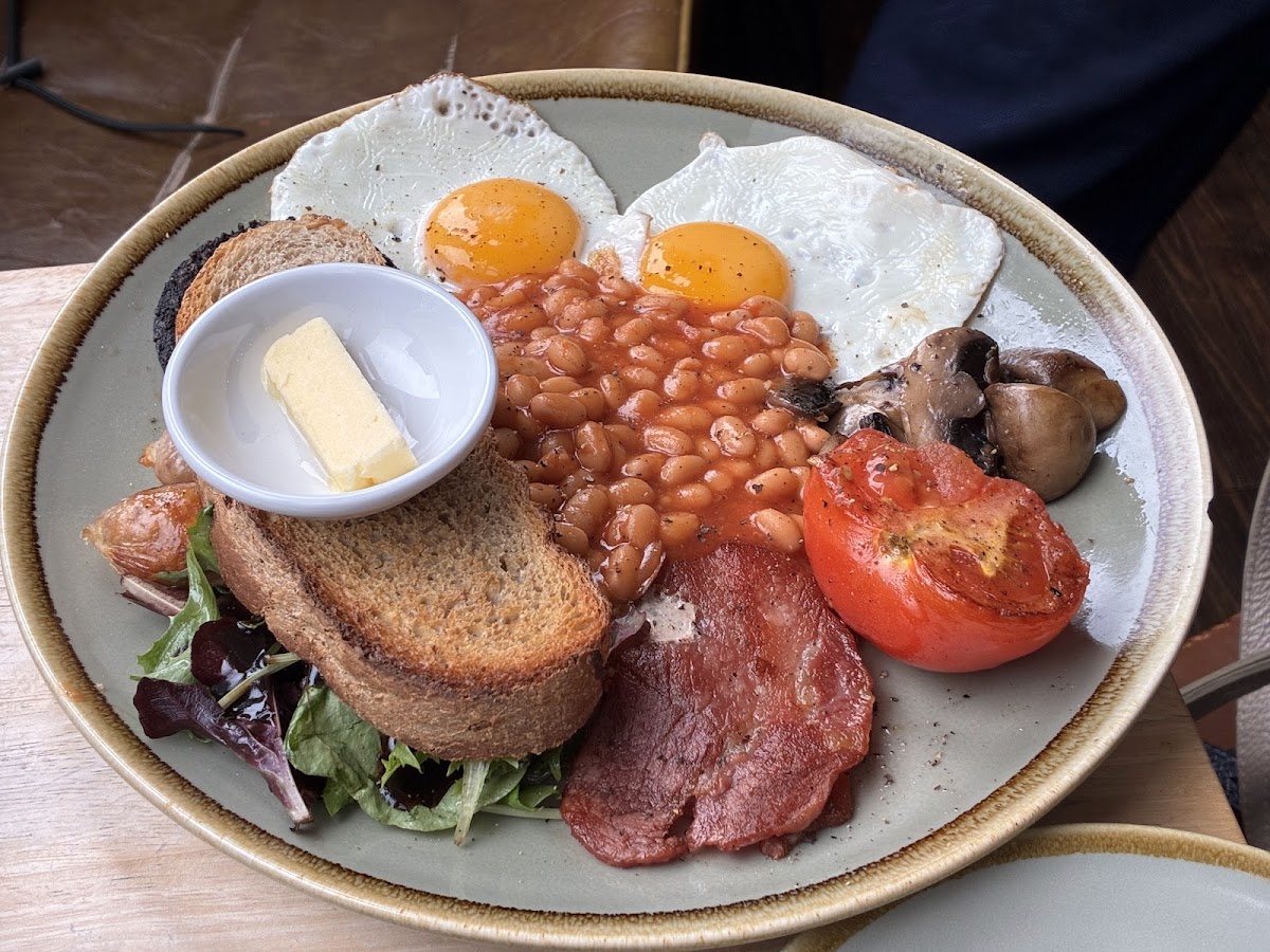 A full breakfast: two fried eggs, beans, bacon, a grilled tomato, mushrooms, black pudding and brown toast with butter.